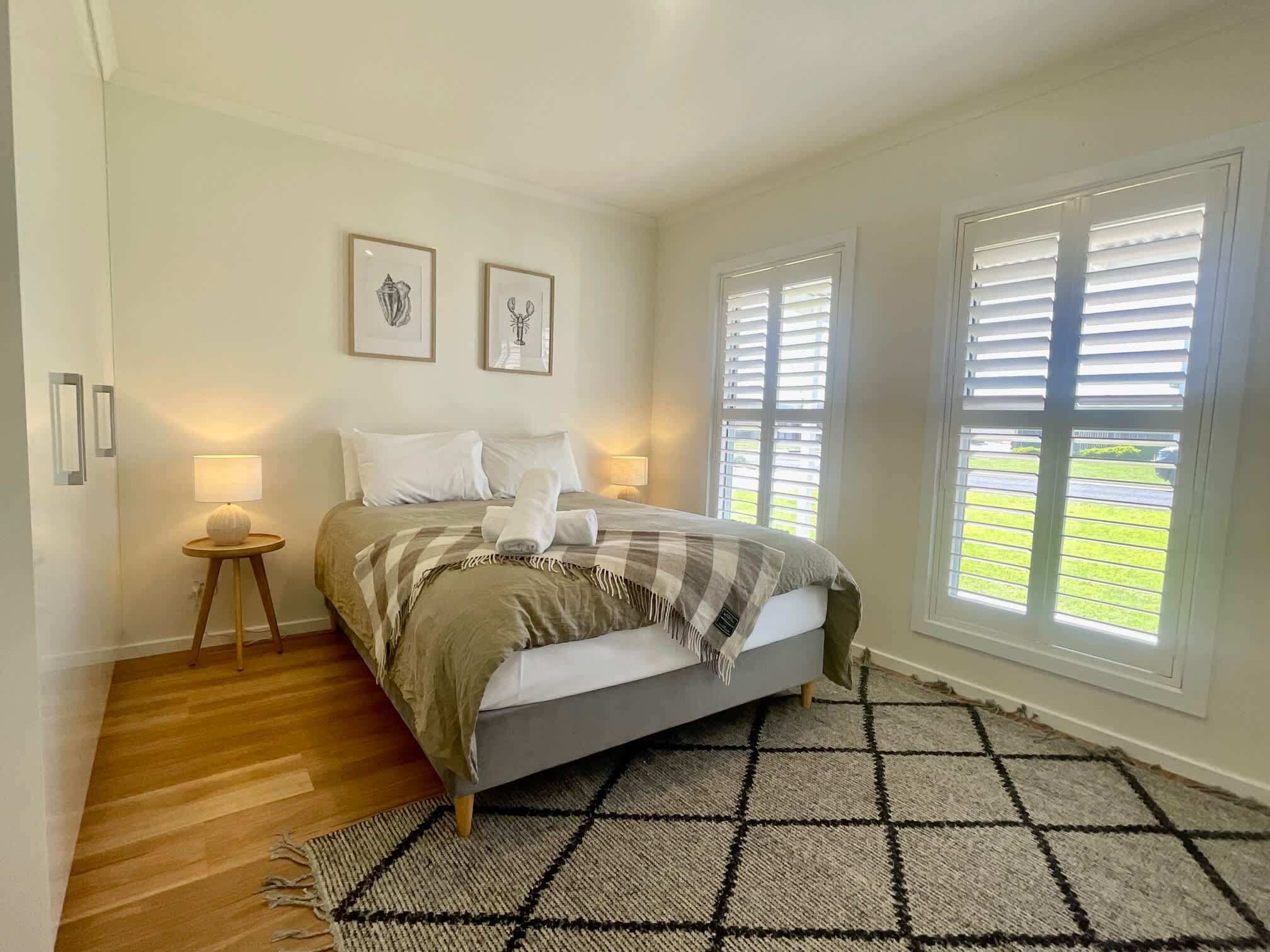 A bedroom with a bed, two bedside lamps, a window with blinds, and a view of a green lawn.