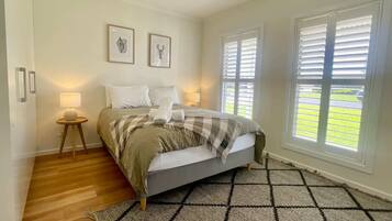 A bedroom with a bed, two bedside lamps, a window with blinds, and a view of a green lawn.