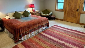 Um quarto com cama, almofadas, mesa de cabeceira, abajur, cadeira, tapete e porta de madeira.