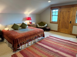 Un dormitorio con cama, almohadas, una mesita de noche, lámpara, silla, alfombra y puerta de madera.