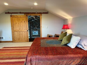 A bedroom with a large bed, a wooden door, a red lamp, and a view of the outdoors.