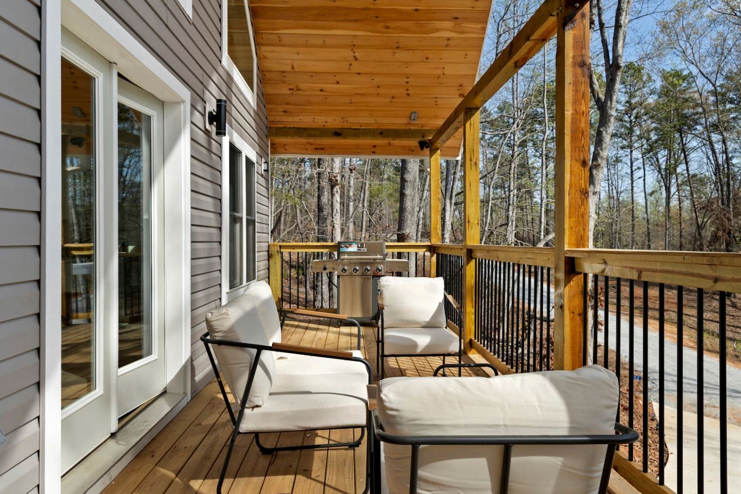 A wooden deck with two white cushioned chairs and a grill, overlooking a forested area.