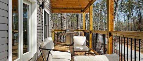 A wooden deck with two white cushioned chairs and a grill, overlooking a forested area.