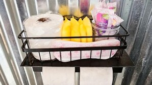 Ein Metallregal mit einem weißen Handtuch, einem gelben Gegenstand und einem pinkfarbenen Behälter vor einer welligen Metallwand.