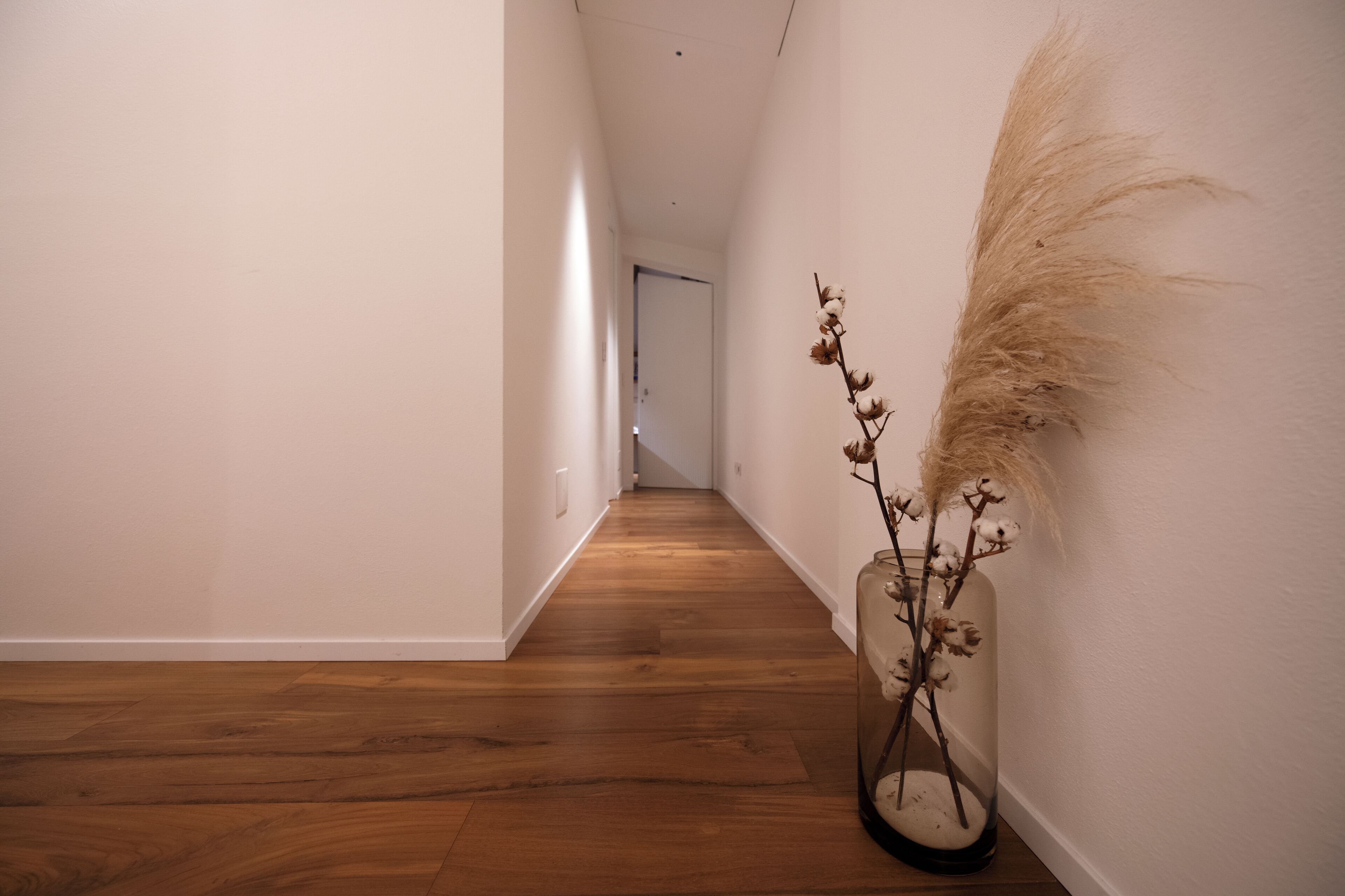 Un couloir au sol en bois, avec un vase contenant des plantes séchées sur le mur de droite.
