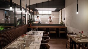 Ein Restaurant mit einem langen Holztisch, gedeckt für das Essen, einem Barbereich und einer durch eine Glasscheibe einsehbarer Küche.