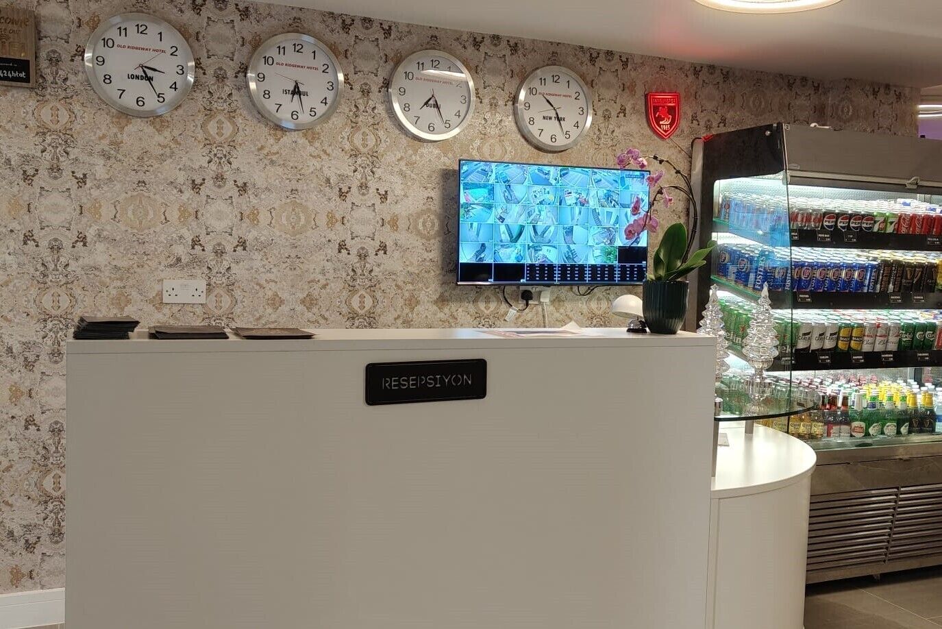 Un'area reception di un hotel con una scrivania reception con la scritta "RESEPSIYON", diverse sveglie appese al muro, uno schermo televisivo che mostra un evento sportivo e un frigorifero pieno di bevande.