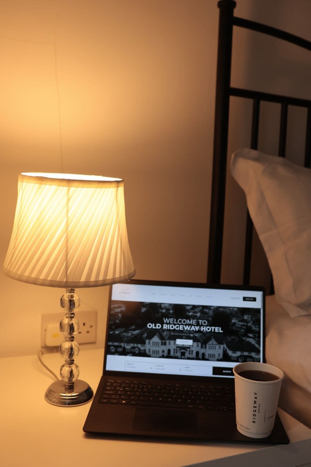 A laptop on a bedside table with a lamp, a cup, and a pillow.