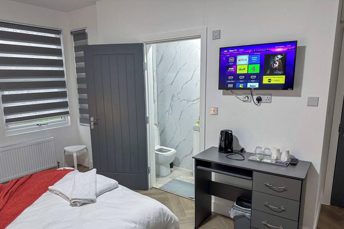 A modern hotel room with a bed, a desk, a TV mounted on the wall, and a bathroom visible through an open door.