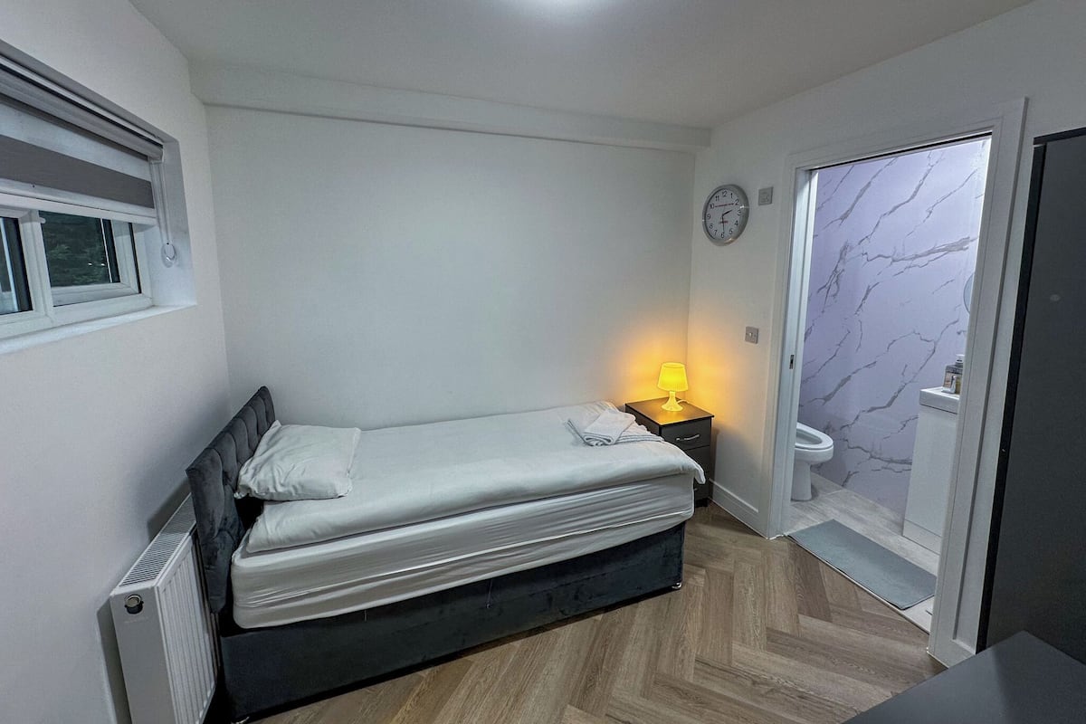 A small, modern bedroom with a bed, bedside lamp, and a view of the bathroom through an open door.