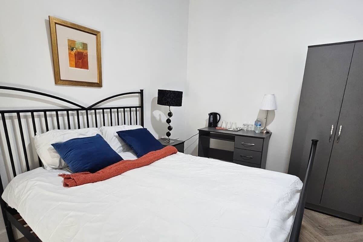 A neatly made bed with white bedding and blue and red pillows, a bedside table with a lamp, a coffee maker, and a wardrobe.