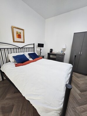 A neatly made bed with white bedding and blue and red pillows, a bedside table with a lamp, a coffee maker, and a wardrobe.