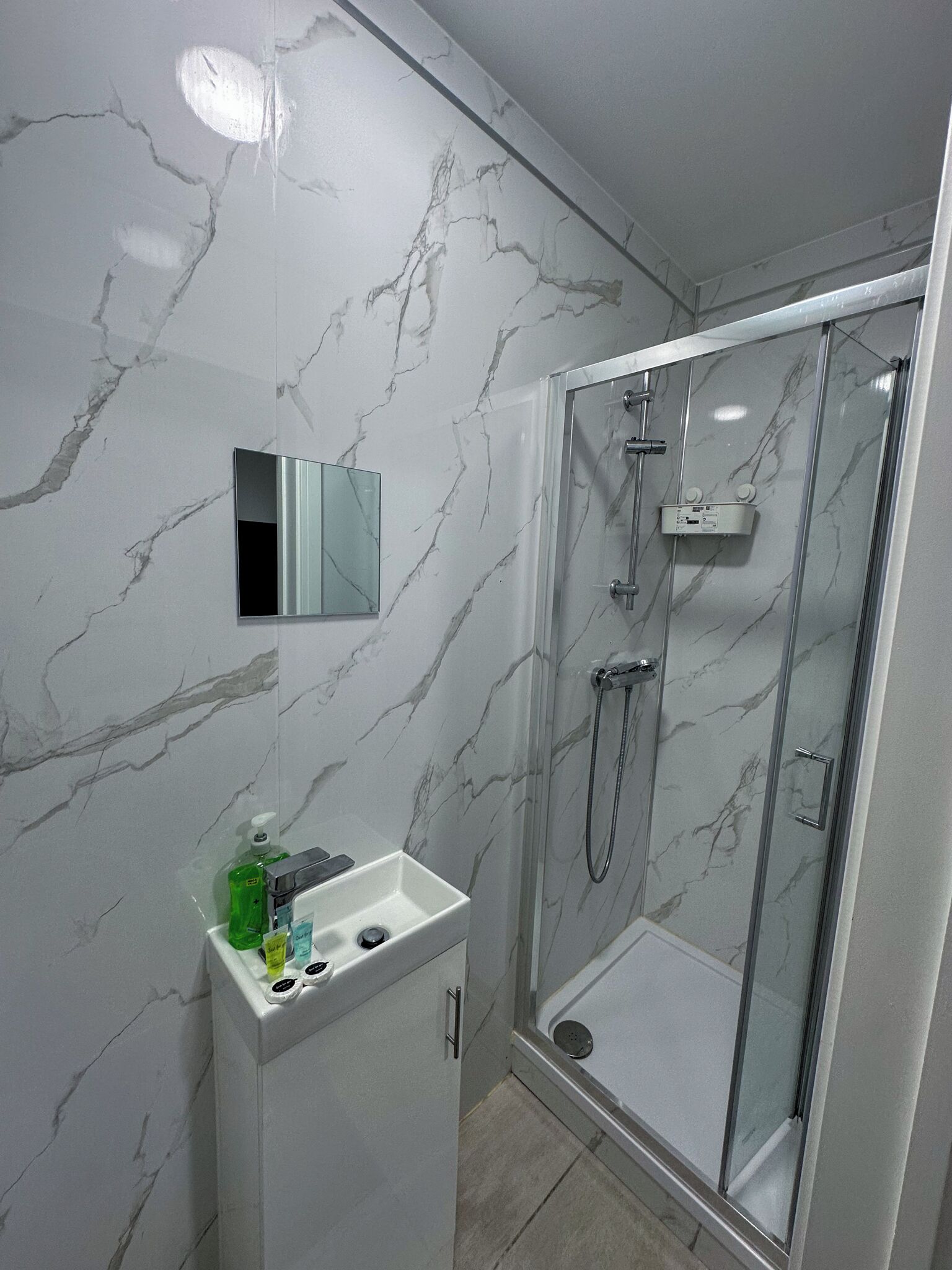 A modern bathroom with a marble wall, a glass shower enclosure, and a vanity with a mirror and toiletries.