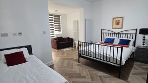 Hotellihuone, jossa on kaksi sänkyä, puinen kalanruotokuvioinen lattia, valkoiset seinät, kattovalaisin, kehystetty taulu ja yöpöytä, jossa on lamppu.