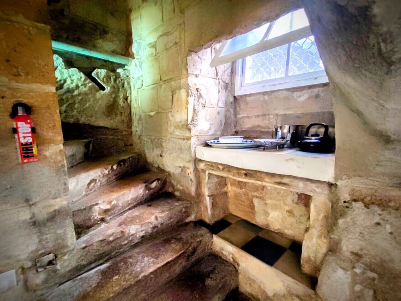 Un interior rústico de piedra con una escalera de madera, una ventana pequeña y una zona de lavamanos.