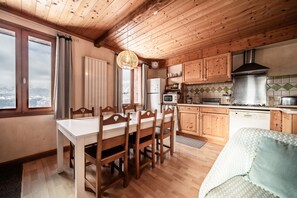 A wooden-paneled kitchen with a dining table and chairs, a sofa, and a view of snowy mountains through large windows.