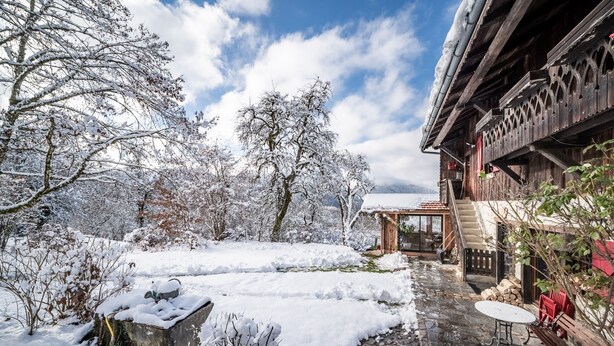 Ein verschneiter Innenhof mit einem Holzgebäude, einem Steinweg und einer Sitzgelegenheit im Freien.