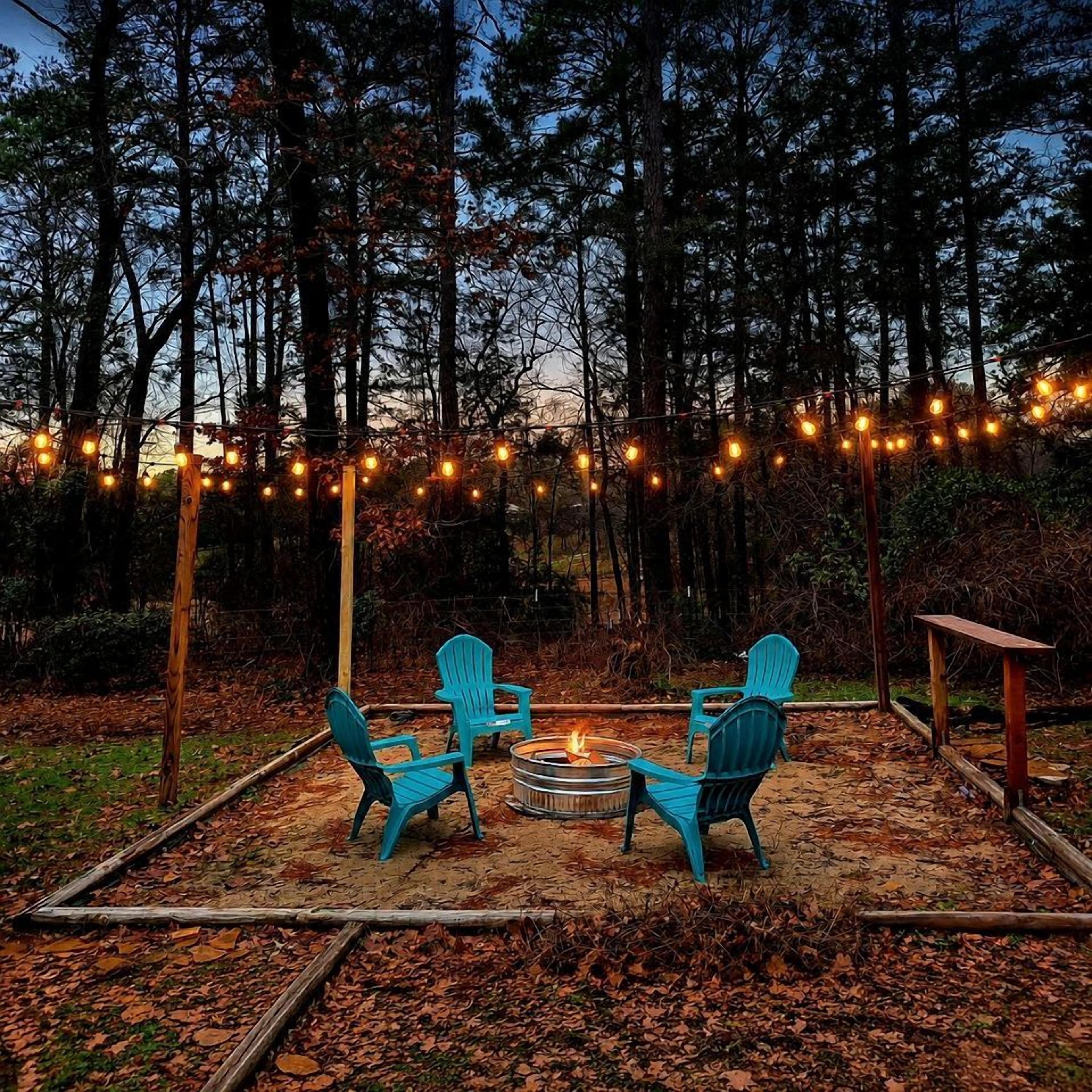An outdoor seating area with four chairs arranged around a central fire pit, surrounded by a forested backdrop and string lights.