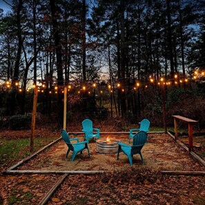Un espace extérieur aménagé avec des sièges, comprenant quatre chaises disposées autour d’un foyer central, le tout entouré d’une forêt et illuminé par des guirlandes lumineuses.