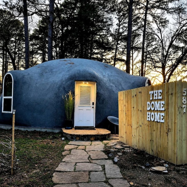 Een koepelvormige constructie met een deur en een houten omheining, waarop 'THE DOME HOME' staat.