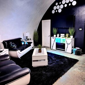 A modern living room with a black leather sofa, a white coffee table, and a black shag rug. There is a white door and a reception desk with plants.