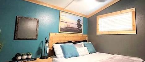 Un dormitorio con cama, mesita de noche, lámpara, ventana con persianas, un cuadro en la pared y ventilador de techo.