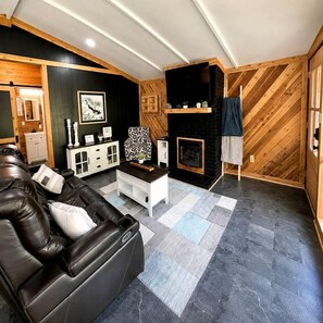 A modern living room with a leather sofa, a fireplace, and a herringbone-patterned wall. - Fish Cabin at the Pines: Cozy Cabin near Lake O' the Pines in Jefferson, Texas (Jefferson)