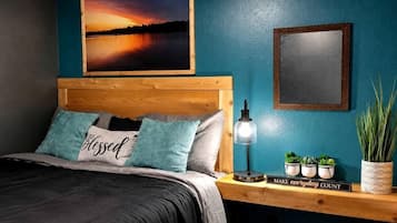 A bedroom with a bed, a nightstand with a lamp, a framed picture, and a potted plant.