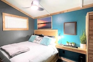 A bedroom with a bed, a nightstand, a lamp, a framed picture, and a window with blinds.