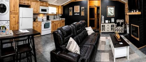 Un espace de vie compact comprenant une cuisine, un coin repas et un coin salon, avec un ventilateur de plafond, des meubles en bois et une cheminée.