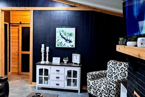 A room with a dark wall, a white cabinet with drawers, a patterned armchair, a framed picture, and a wooden shelf with decorative items.