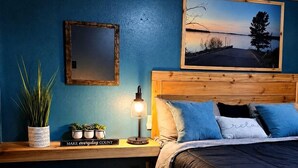 A bedroom with a wooden headboard, a bed with dark bedding, a nightstand with a lamp, a framed picture on the wall, and a small potted plant. - Fish Cabin at the Pines: Cozy Cabin near Lake O' the Pines in Jefferson, Texas (Jefferson)