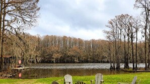 A grassy area with several wooden Adirondack chairs arranged in a semi-circle, overlooking a calm lake with trees in the background.