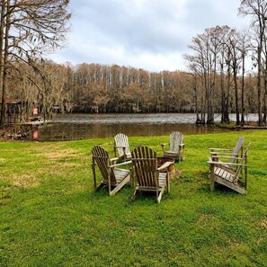A grassy area with several wooden Adirondack chairs arranged in a semi-circle, overlooking a calm lake with trees in the background. - Keepin It Reel on Caddo Lake: Incredible Houseboat in Uncertain, Texas (Uncertain)