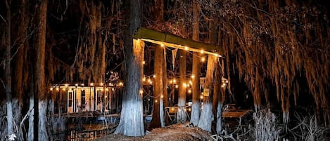 A rustic wooden structure with string lights, surrounded by trees and a path leading to it.