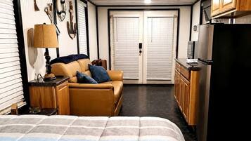 A compact hotel room with a bed, a sofa, a small table with a lamp, a kitchenette, and a door leading to another room.