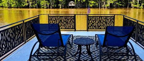 Terraza con dos sillas y una pequeña mesa, con vista a un cuerpo de agua y árboles al fondo.
