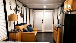 A compact hotel room with a bed, a sofa, a small table with a lamp, a kitchenette, and a door leading to another room.