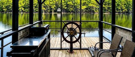 A covered deck with a wooden floor, a steering wheel, and two chairs.