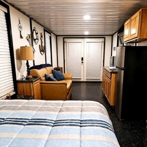A compact hotel room with a bed, a sofa, a small table with a lamp, a kitchenette, and a door leading to another room. - Keepin It Reel on Caddo Lake: Incredible Houseboat in Uncertain, Texas (Uncertain)