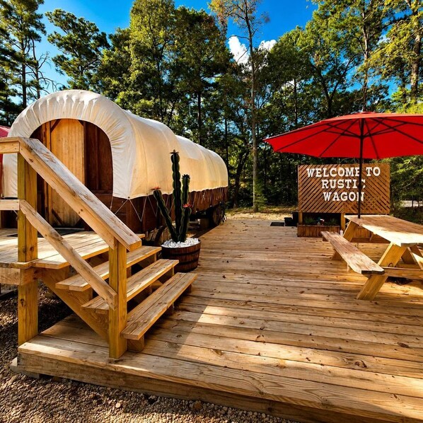 A rustic wagon with a covered sleeping area, wooden steps leading up to it, a red umbrella, and a welcome sign.