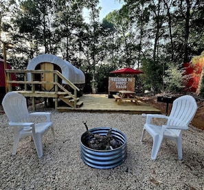 Un espacio de estar al aire libre rústico con sillas Adirondack, un pozo de fuego, un escenario de madera y un cartel de bienvenida.