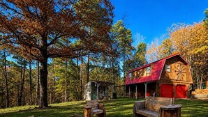 A rustic cabin with a red roof, surrounded by a grassy yard with wooden furniture and a metal fire pit. - Rustic Retreat near Lake O the Pines: Gorgeous Barn in Warlock, Texas (Warlock)