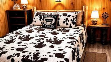 Een slaapkamer met een bedovertrek met koeienprint, een kussen met de tekst 'MEADOW FARMS', een houten hoofdbord, twee bedlampen en een nachtkastje van hout met een windmolen decoratie.