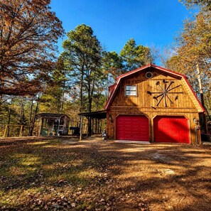 A wooden barn with a red garage door, surrounded by trees and a covered structure.