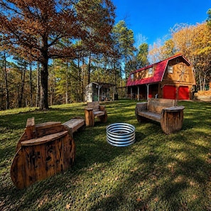A rustic cabin with a red roof, surrounded by a grassy yard with wooden furniture and a metal fire pit. - Rustic Retreat near Lake O the Pines: Gorgeous Barn in Warlock, Texas (Warlock)