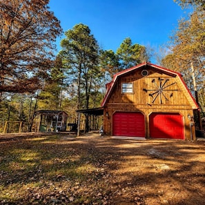 A wooden barn with a red garage door, surrounded by trees and a covered structure. - Rustic Retreat near Lake O the Pines: Gorgeous Barn in Warlock, Texas (Warlock)