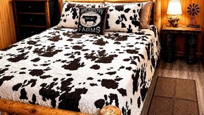 A bedroom with a cow print bedspread, a pillow with 'MEADOW FARMS' text, wooden headboard, two bedside lamps, and a wooden nightstand with a windmill decoration. - Rustic Retreat near Lake O the Pines: Gorgeous Barn in Warlock, Texas (Warlock)