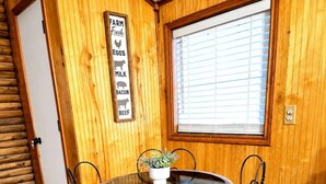 Una habitación con paneles de madera, una mesa redonda y cuatro sillas, una ventana con persianas y un cartel en la pared que enumera productos agrícolas.