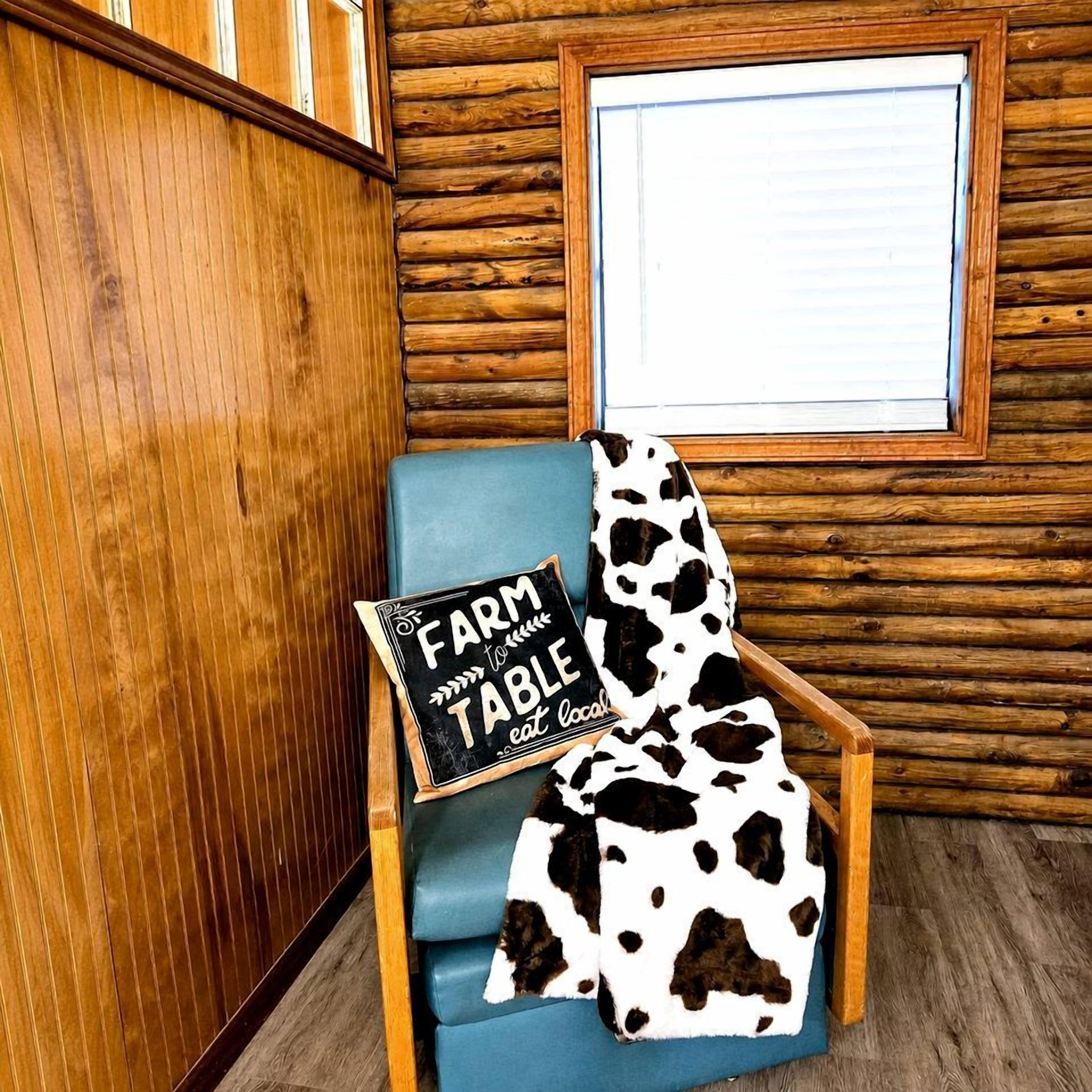 A rustic wooden cabin interior with a blue cushioned chair, a cow print blanket, and a sign reading 'FARM TABLE'.